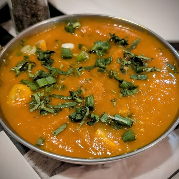A close-up of a vibrant orange Indian curry, garnished with fresh green herbs in a metal bowl.
