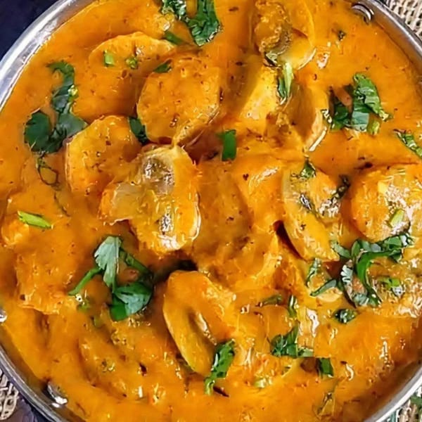 Close-up of a vibrant orange Indian mushroom curry, garnished with fresh green cilantro in a metal bowl.